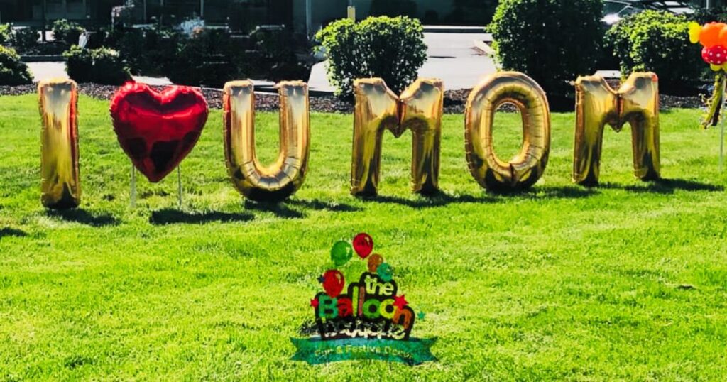Outdoor yard balloon decor spelling out 'I Love You Mom' welcomes guests to a Mother's Day celebration, creating a festive and inviting atmosphere.