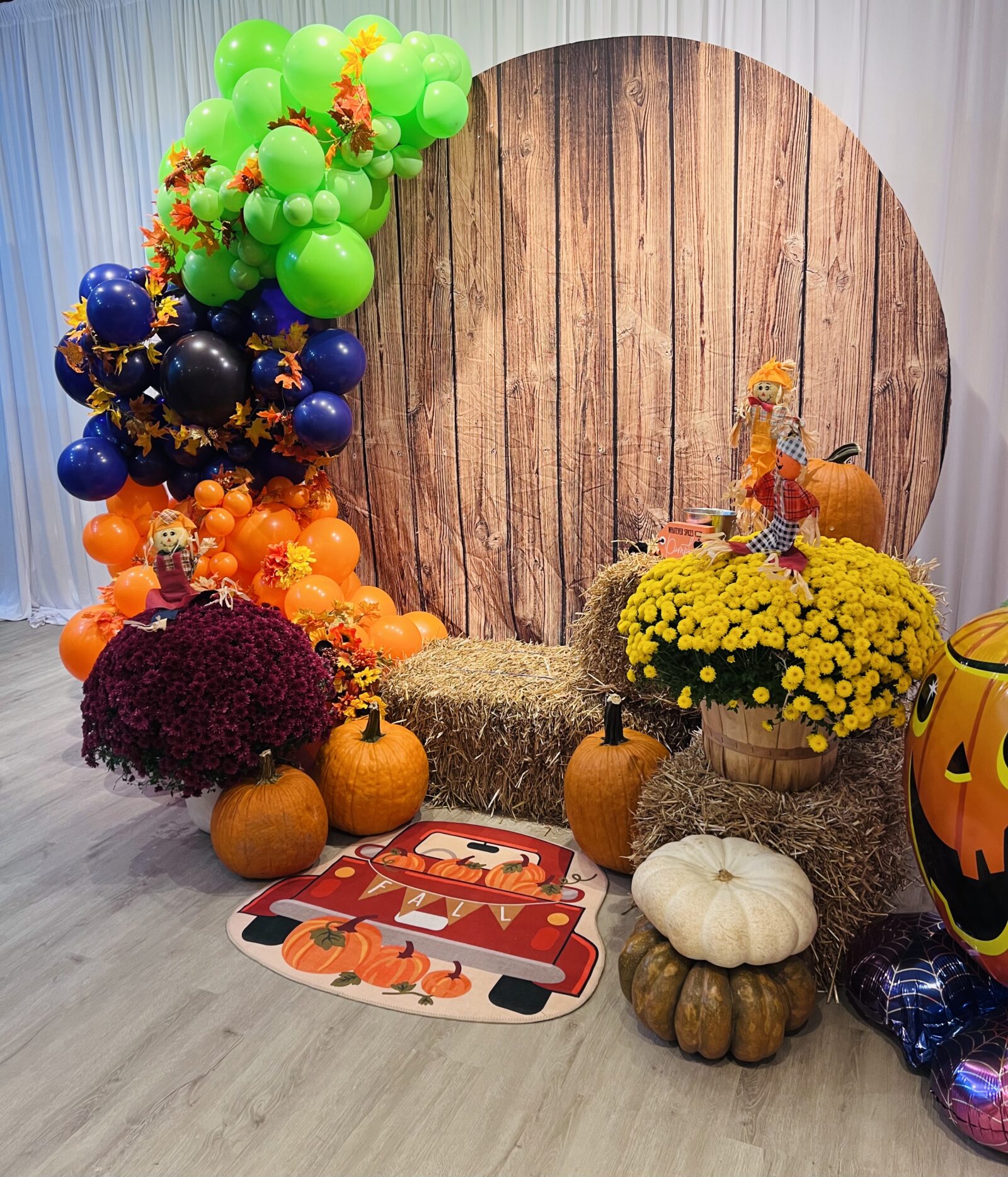 Organic balloon garland in orange, purple, and lime green balloons forming a festive selfie station, accented with hay, pumpkins, and a colorful rug.