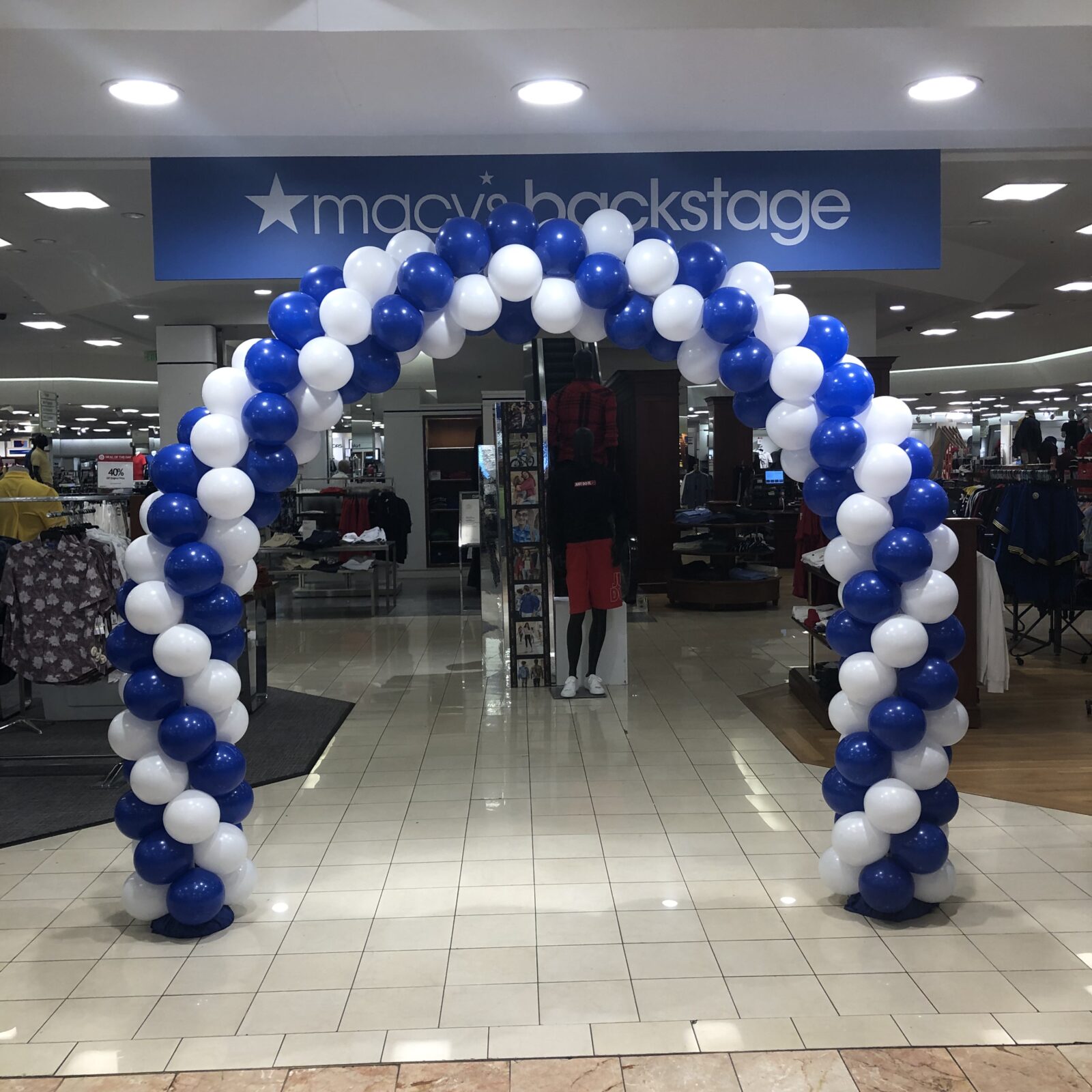 A balloon arch made of blue and white balloons in a classic spiral pattern stands at the entrance of a Macy's Backstage store inside a shopping mall. The arch frames the entrance, creating a welcoming and festive atmosphere. The surrounding area features clothing racks, mannequins, and store displays.